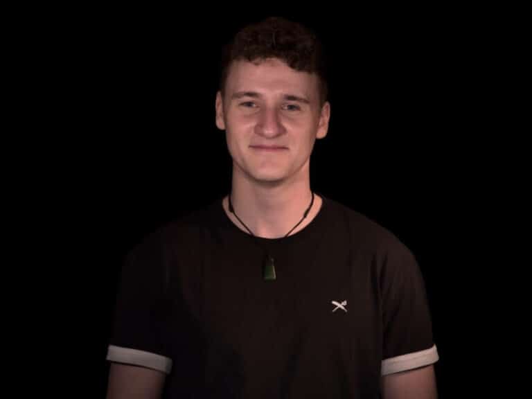 Ein Porträt von DJ Mambl mit schwarzem T-Shirt. Er hat eine braune Kette um den Hals und wird vor einem dunklen Hintergrund beleuchtet.